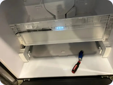 Open refrigerator ice-maker compartment with its cover raised and a hand tool below.