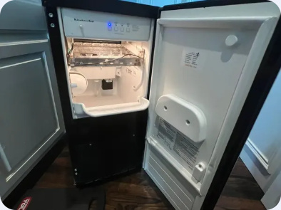 Undercounter ice machine with its door open, revealing the ice-making compartment and storage area.
