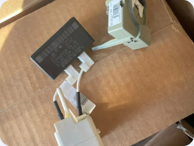 Removed refrigerator start-device components and a capacitor resting on cardboard.