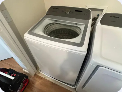 Samsung top-load washing machine with a glass lid and a service tool bag beside it.