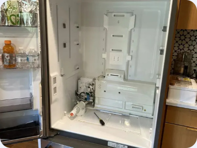 Refrigerator interior with shelves and panels removed, exposing lower internal components and a hand tool.